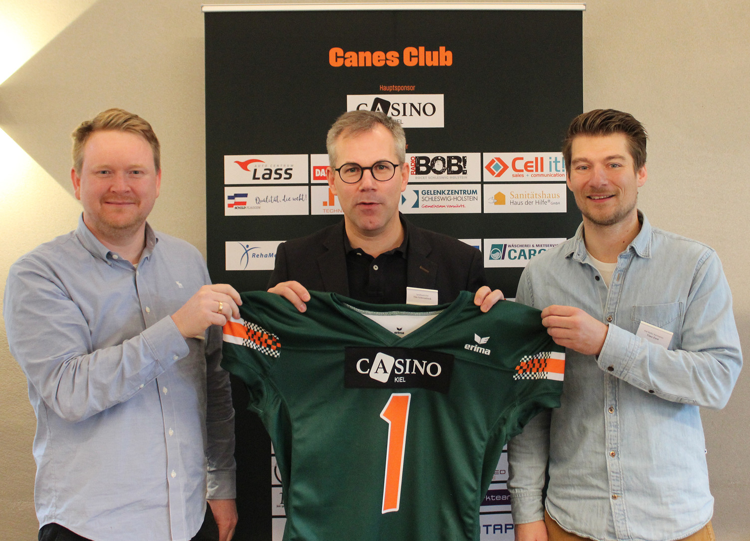 Michael Herrmann (links) und Timo Zorn (rechts) von den Hurricanes halten zusammen mit Tilo Schwonbeck (Mitte), Direktor des Casino Kiel, ein grünes Hurricanes-Trikot mit der Nummer 1 hoch. Über der Eins prangt das Logo des Casino auf der Brust. 